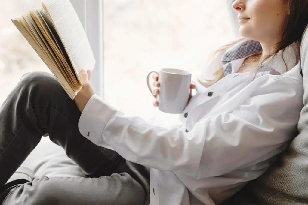 woman sitting by a window reading a manifestation book while drinking coffee